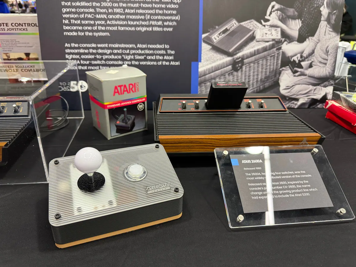 The silver Arkade controller with a white ball-top joystick, on a display table next to a wood-grain Atari 2600A and a boxed Atari joystick.