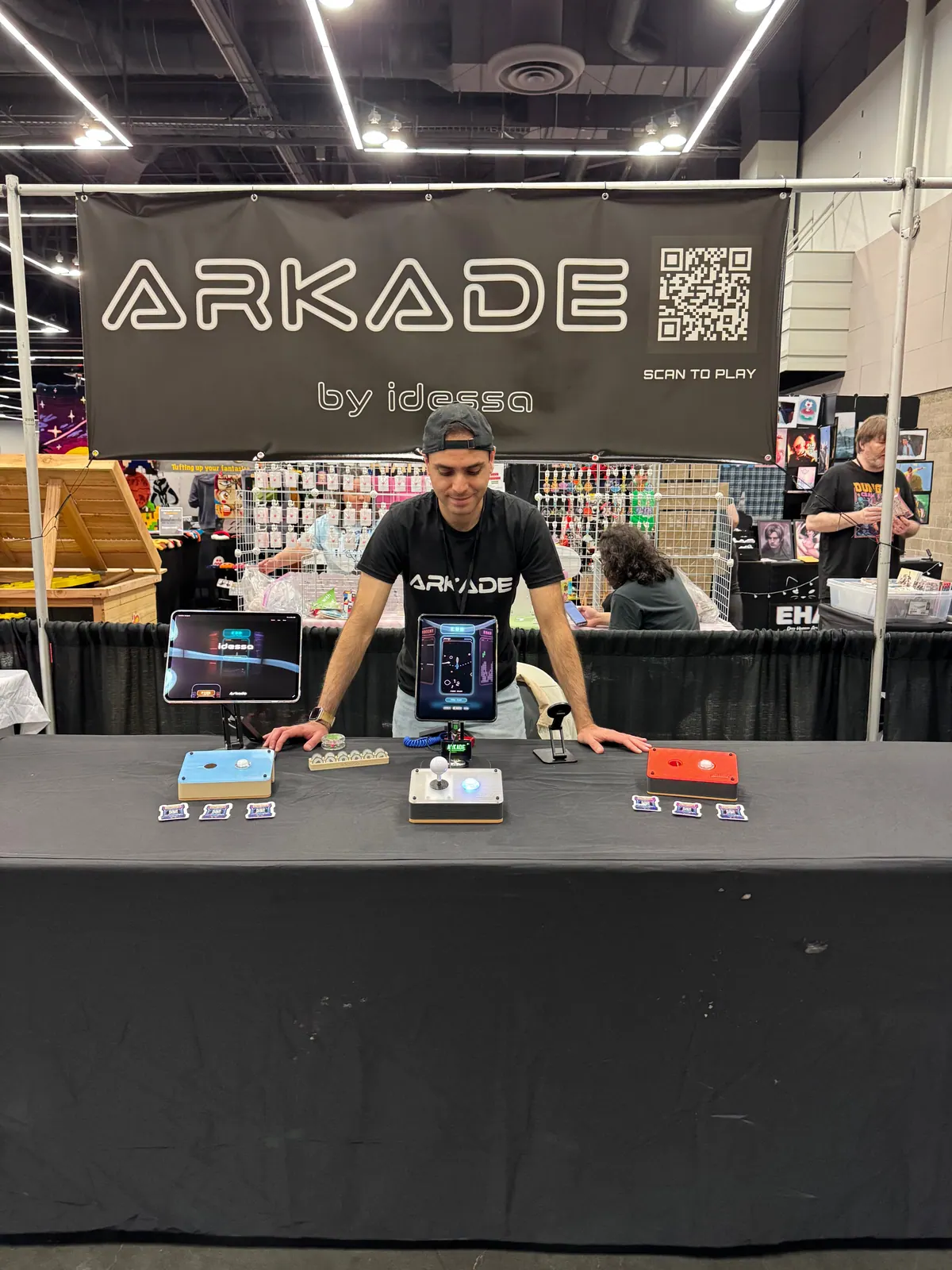 Albert behind a black table set with arcade controllers and iPads, under a banner that reads Arkade by idessa, scan to play.