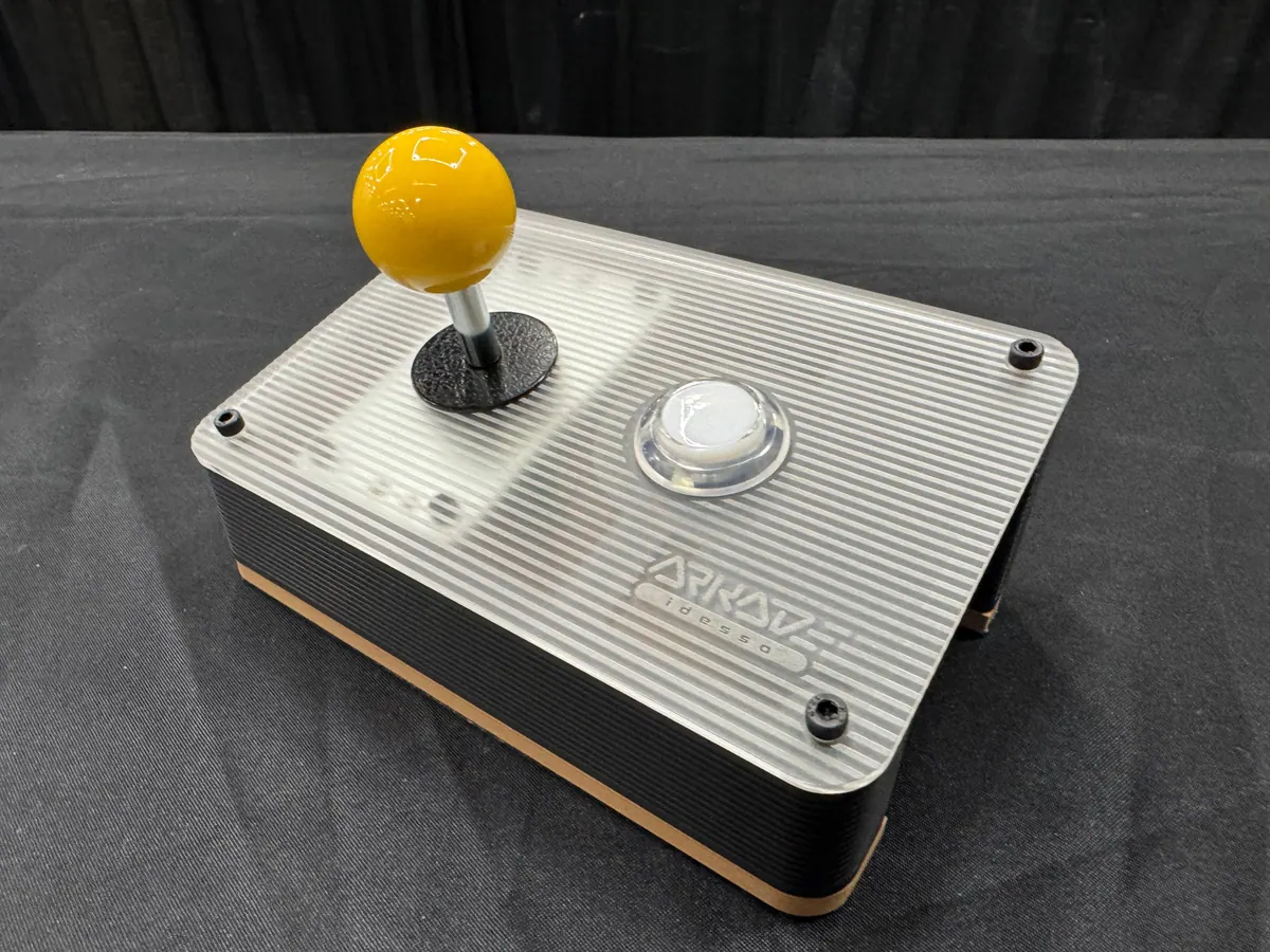 The Arkade controller: a ribbed silver top with a yellow ball-top joystick and a white button, on a black base with a copper stripe.