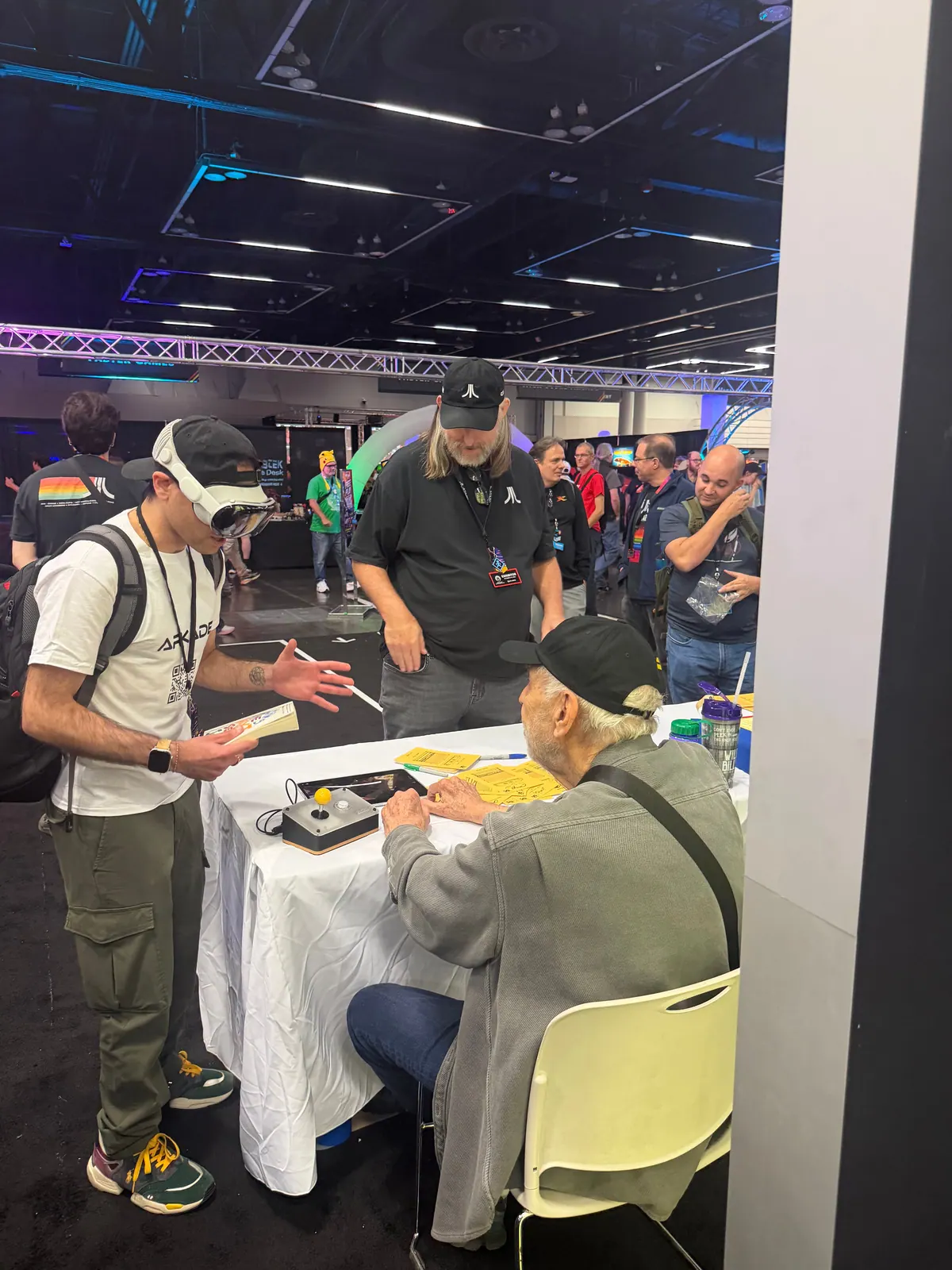 Albert, wearing an Apple Vision Pro, at a table where Nolan Bushnell sits with the Arkade controller in front of him, a crowd looking on.