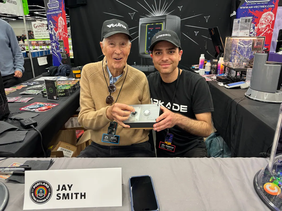 Jay Smith, in a Vectrex cap, holding the Arkade controller beside Albert at his signing table, behind a card that reads Jay Smith.